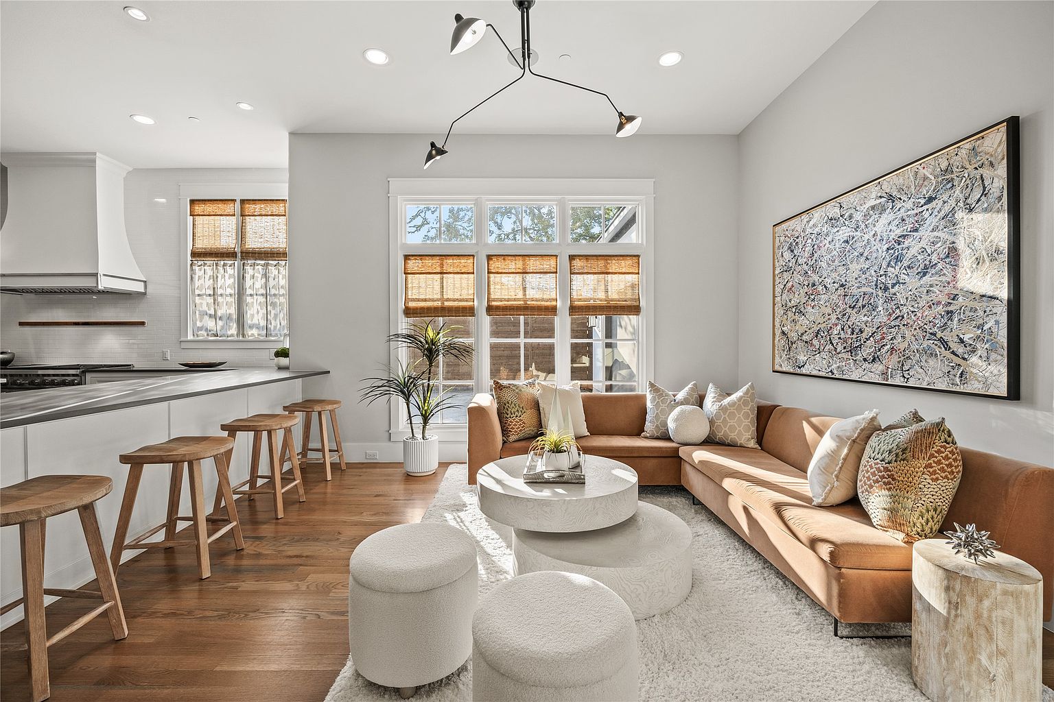 This is an interior shot of a modern living room featuring a sectional sofa, layered round coffee tables, and a large abstract art piece. The room is well-lit with natural light from a large window and features hardwood floors and neutral tones, creating a warm and inviting atmosphere. The perspective is from a medium distance, capturing the entire living space and its connection to the adjacent kitchen area.