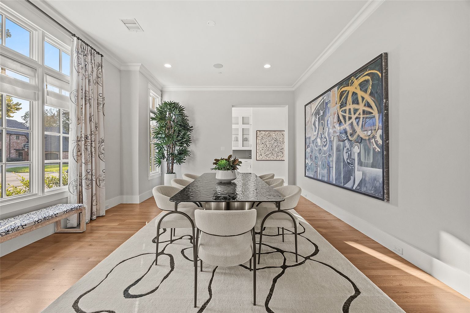 This is an interior shot of a dining room featuring a modern aesthetic. The room includes a long, dark dining table surrounded by light-colored chairs, set atop a large, patterned rug. A large piece of abstract art hangs on the wall, and natural light floods the space through large windows, complemented by a tall potted plant in the corner.