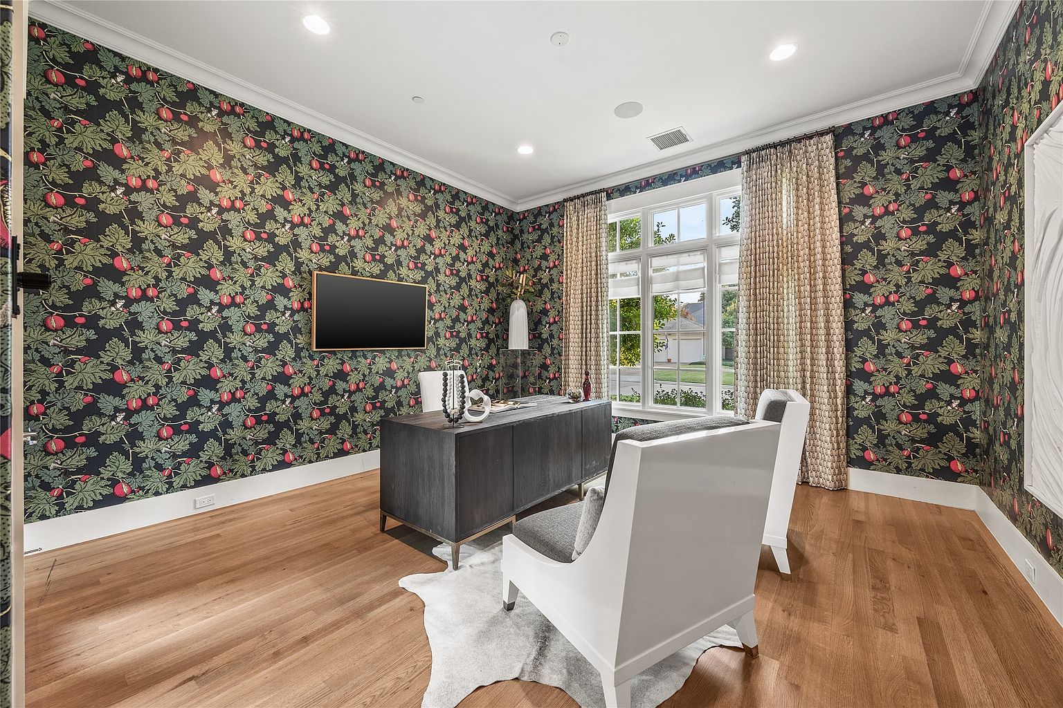 This is an interior shot of a home office featuring dark floral wallpaper, hardwood floors, and white trim. A dark wood desk sits in the center of the room, flanked by two white chairs with gray cushions. A large window provides natural light, and a flat-screen TV is mounted on the wall, creating a sophisticated and functional workspace.