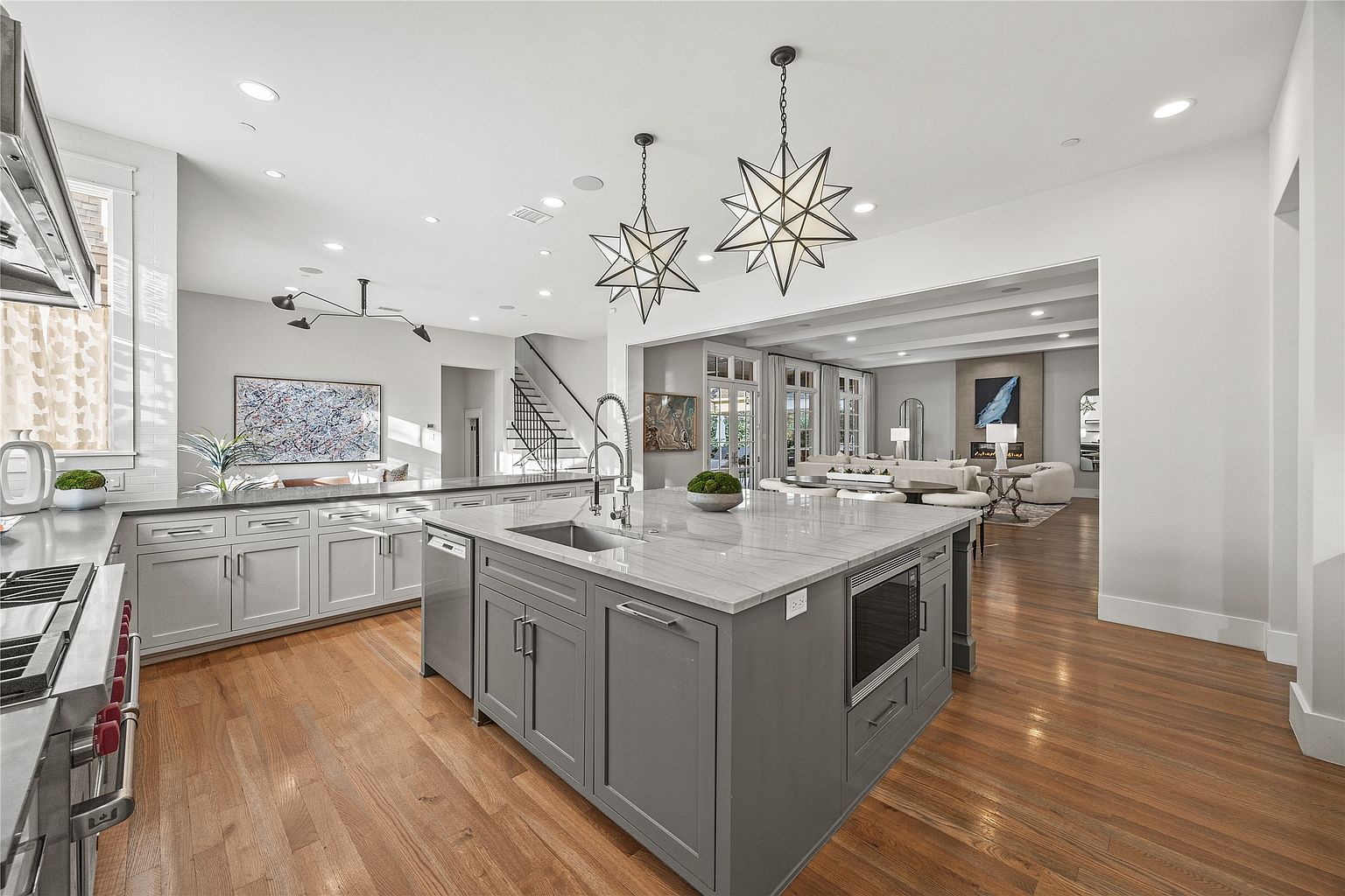 This is a bright and spacious kitchen featuring a large central island with a sink and microwave, gray cabinetry, and hardwood floors. Star-shaped pendant lights hang above the island, adding a touch of modern elegance. The kitchen seamlessly flows into the living area, creating an open and inviting space.