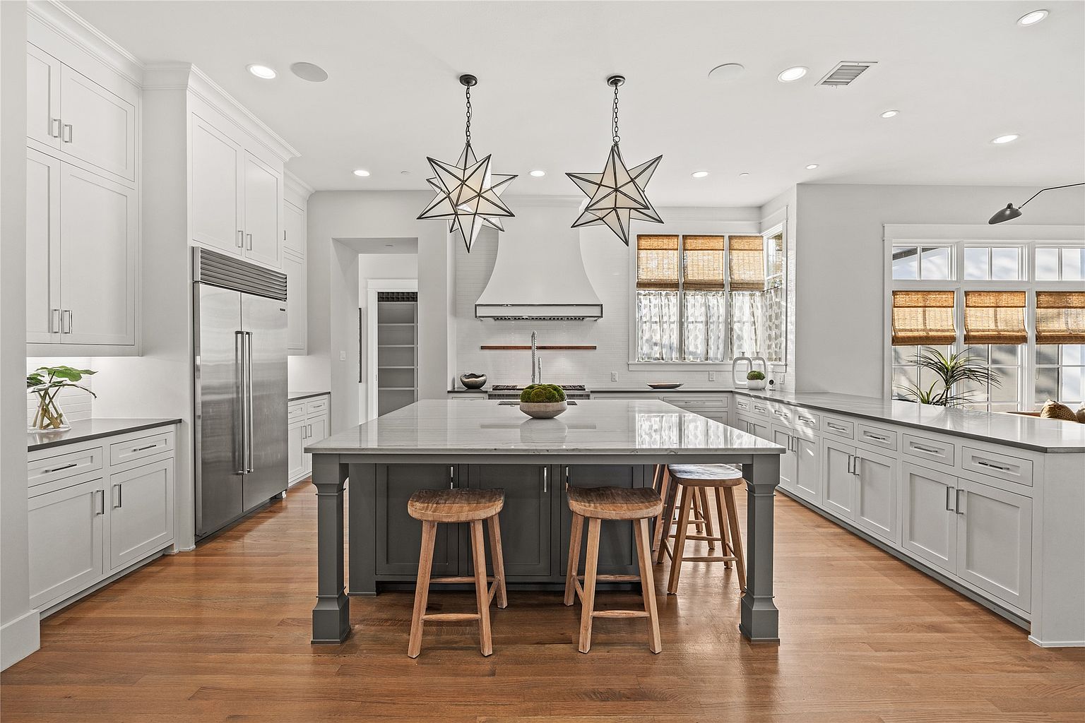 This is a bright and spacious kitchen featuring white cabinetry, stainless steel appliances, and a large gray island with a marble countertop. Two star-shaped pendant lights hang above the island, and natural light floods the space through the windows. The hardwood floors add warmth to the overall design, creating an inviting and modern culinary space.