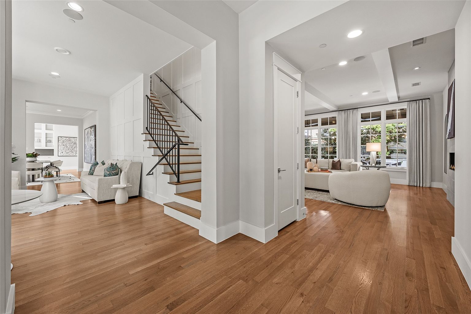 This is an interior shot of a well-lit living room featuring hardwood floors and white walls. The space is divided into two areas, one with a staircase and a seating area with a sofa and the other with a larger seating arrangement near large windows. The overall impression is clean, modern, and inviting, highlighting the spaciousness and natural light.