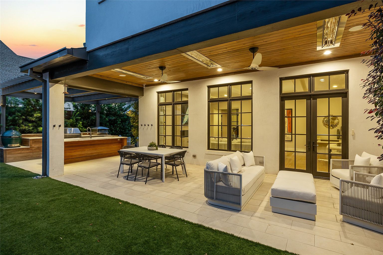 This is a well-appointed outdoor patio area, featuring a dining set, comfortable lounge seating, and an outdoor kitchen. The patio is paved with light-colored tiles and transitions to a well-maintained lawn. The space is covered by a pergola and a roof extension, providing shade and shelter, with ceiling fans and heaters for comfort. The overall impression is one of luxury and relaxation, perfect for outdoor entertaining.