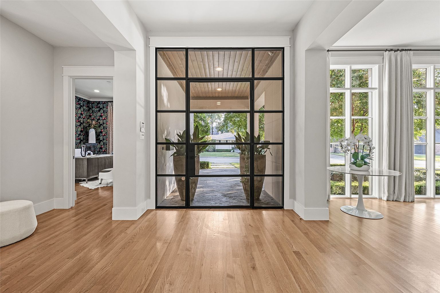 This interior shot showcases a bright and spacious entryway featuring hardwood floors and neutral-toned walls. A large, multi-paned glass door provides a view to the exterior, flanked by potted plants. To the left, an open doorway leads to a room with patterned wallpaper, while to the right, large windows allow natural light to flood the space, complemented by a round table with floral arrangement.