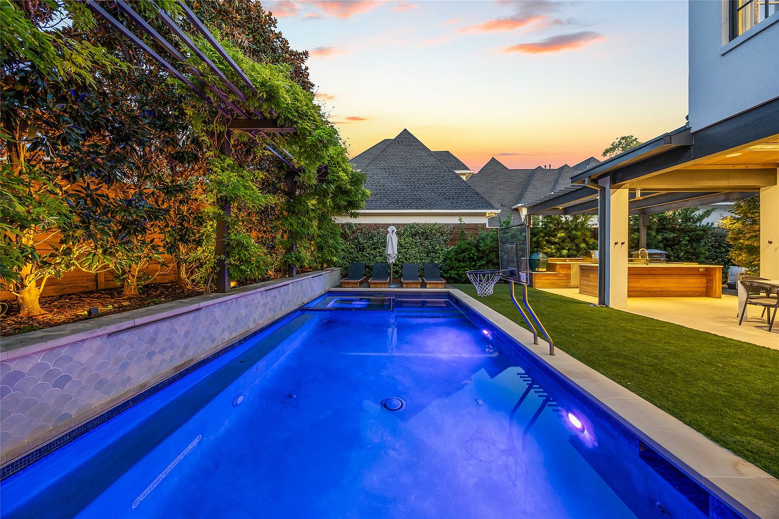 This image showcases a luxurious backyard pool area at dusk. The rectangular pool is illuminated with blue lighting, and lounge chairs are positioned at the far end. A covered outdoor kitchen and dining area are visible to the right, while lush greenery and a pergola frame the space, creating a private and inviting atmosphere.