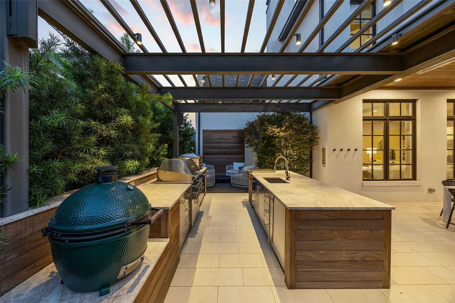 This image showcases a luxurious outdoor kitchen and patio area. The space features a built-in grill, a green egg smoker, and a sink with countertop space, all under a modern pergola. The patio is paved with light-colored tiles, and lush greenery surrounds the area, creating a private and inviting atmosphere.