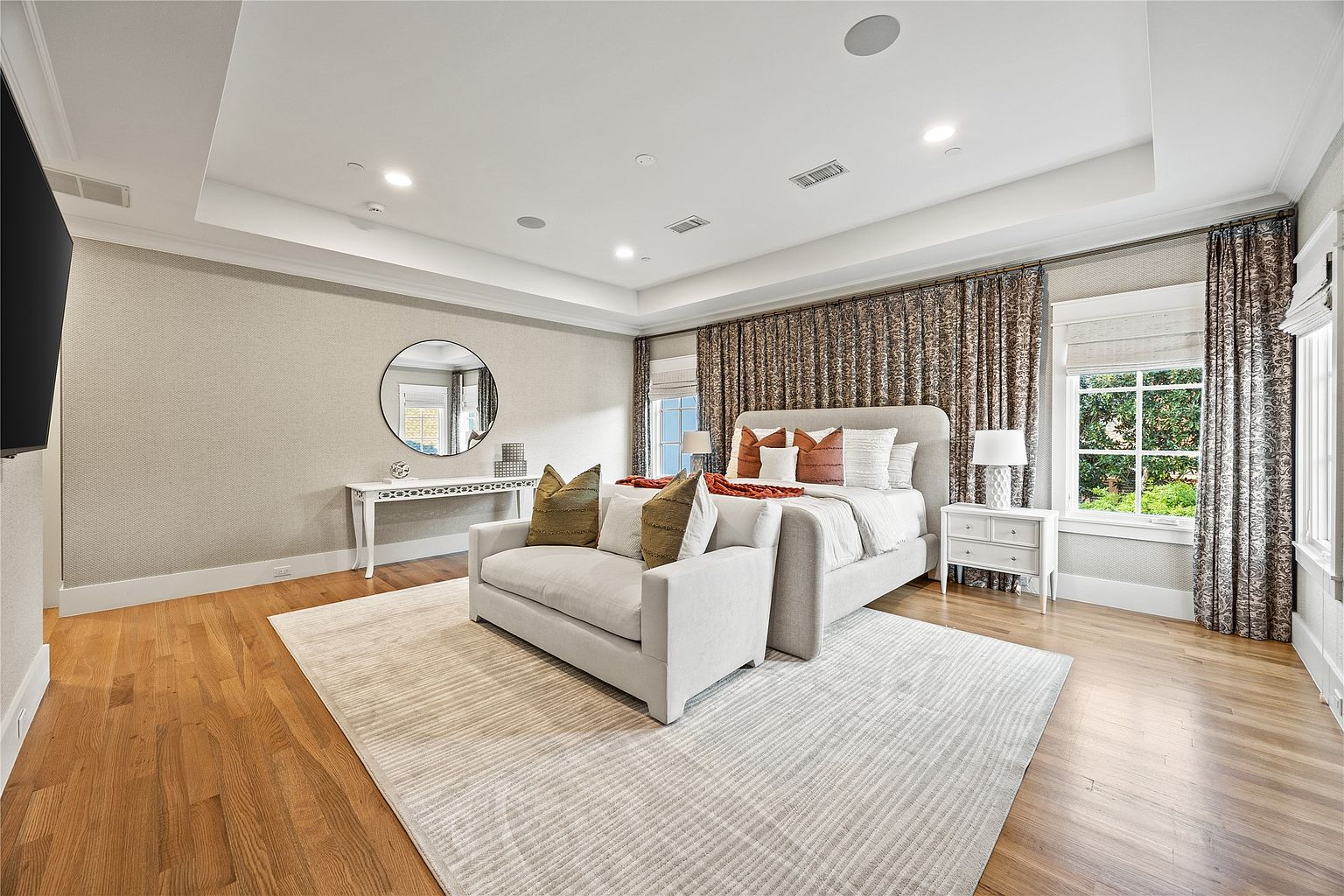 This is a primary bedroom featuring a large bed with a padded headboard, a seating area in front of the bed, and a console table with a round mirror. The room has hardwood floors, a neutral color palette, and patterned curtains that add visual interest. The overall impression is luxurious and comfortable.