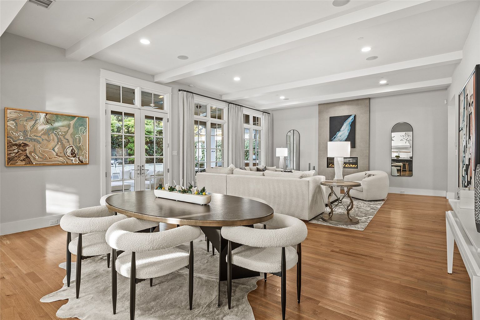 This is an interior shot of a combined living and dining area. The living room features a large sectional sofa, a fireplace with artwork above, and a neutral color palette. The dining area includes an oval table with unique chairs and a cowhide rug, creating a stylish and inviting space with hardwood floors throughout.