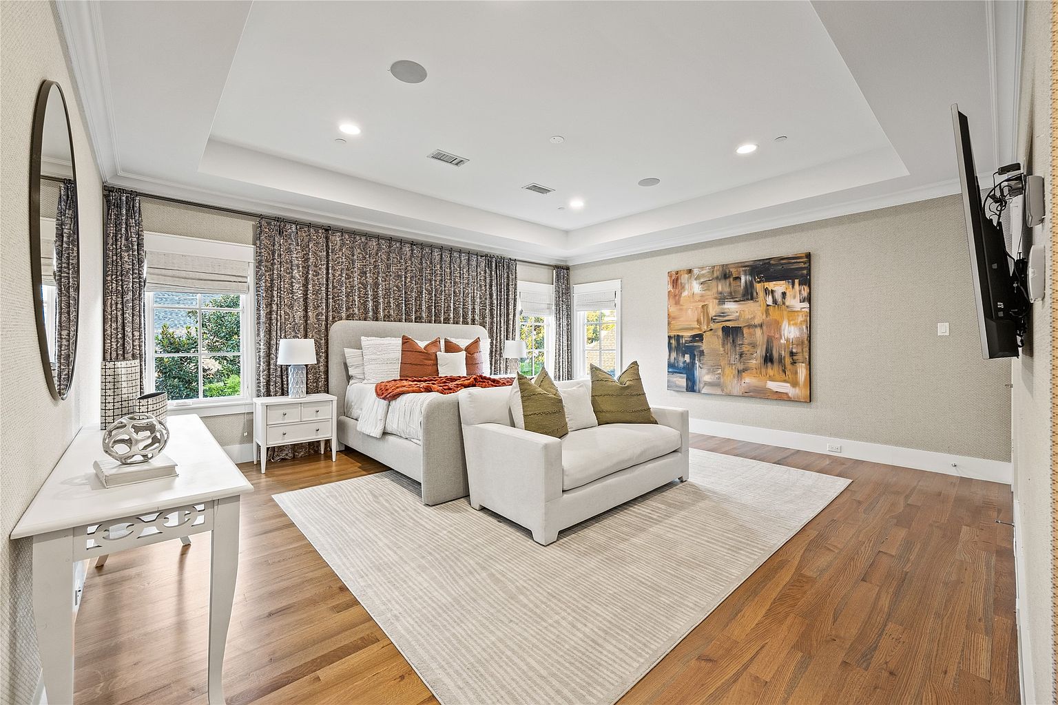 This is a primary bedroom featuring a large bed with decorative pillows and a throw blanket, a seating area with a small sofa, and an abstract painting on the wall. The room has hardwood floors, a neutral-colored rug, and recessed lighting, creating a luxurious and comfortable atmosphere. The curtains add a touch of elegance, and the overall design is sophisticated and inviting.