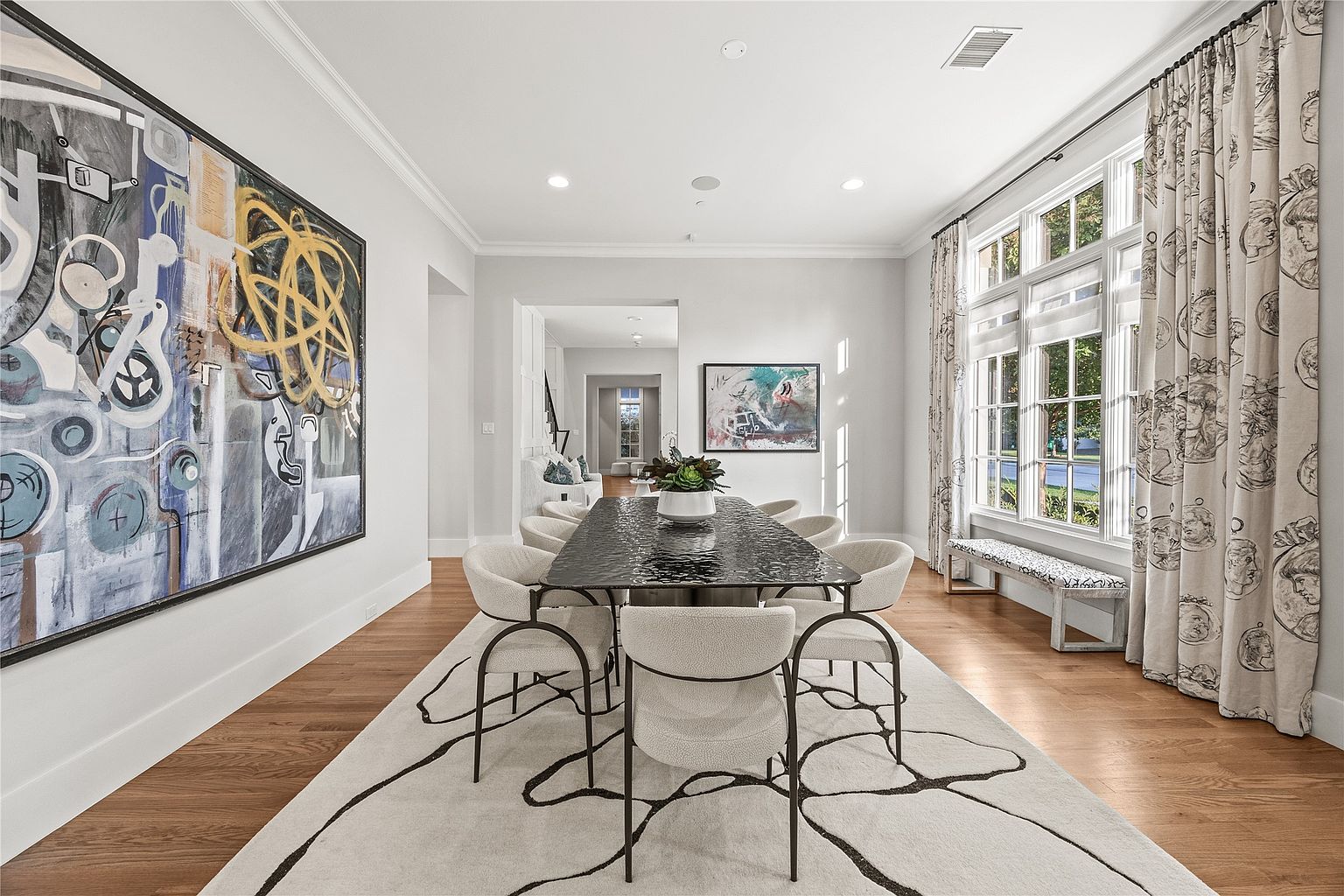 This is an interior shot of a dining room featuring a long, dark-colored dining table surrounded by light-colored chairs with black frames. A large, abstract painting hangs on the left wall, and a window with patterned curtains is on the right, providing natural light. The room is decorated with a modern rug and has hardwood floors, creating an elegant and inviting atmosphere.