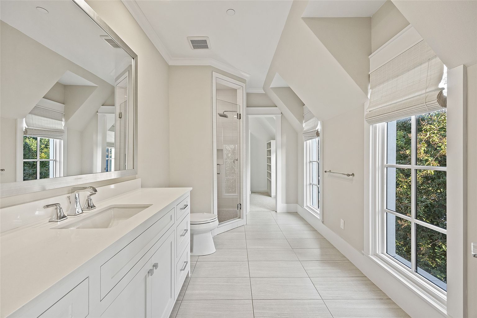 This is a bright and spacious primary bathroom featuring a double vanity with white countertops and cabinetry, a large mirror, and modern chrome fixtures. The bathroom includes a glass-enclosed shower and a toilet, with light gray tile flooring throughout. Natural light floods the room through two large windows with woven shades, creating a clean and serene atmosphere.