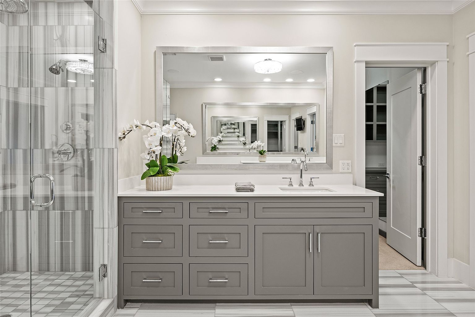 This is a well-lit primary bathroom featuring a gray vanity with multiple drawers and cabinets, topped with a white countertop and a large mirror. The bathroom includes a glass-enclosed shower with gray and white marble-like tiles, and the flooring continues the gray and white theme. The overall impression is clean, modern, and luxurious.