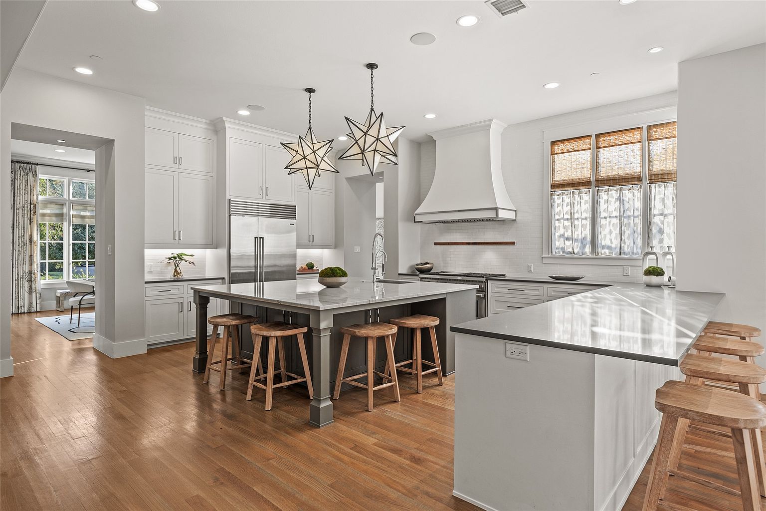 This is a bright and spacious kitchen featuring a large central island with seating, custom cabinetry, and high-end stainless steel appliances. The kitchen is well-lit with recessed lighting and pendant fixtures, and the hardwood floors add warmth to the space. The perspective is from a wide angle, showcasing the entire kitchen layout and its connection to an adjacent room.
