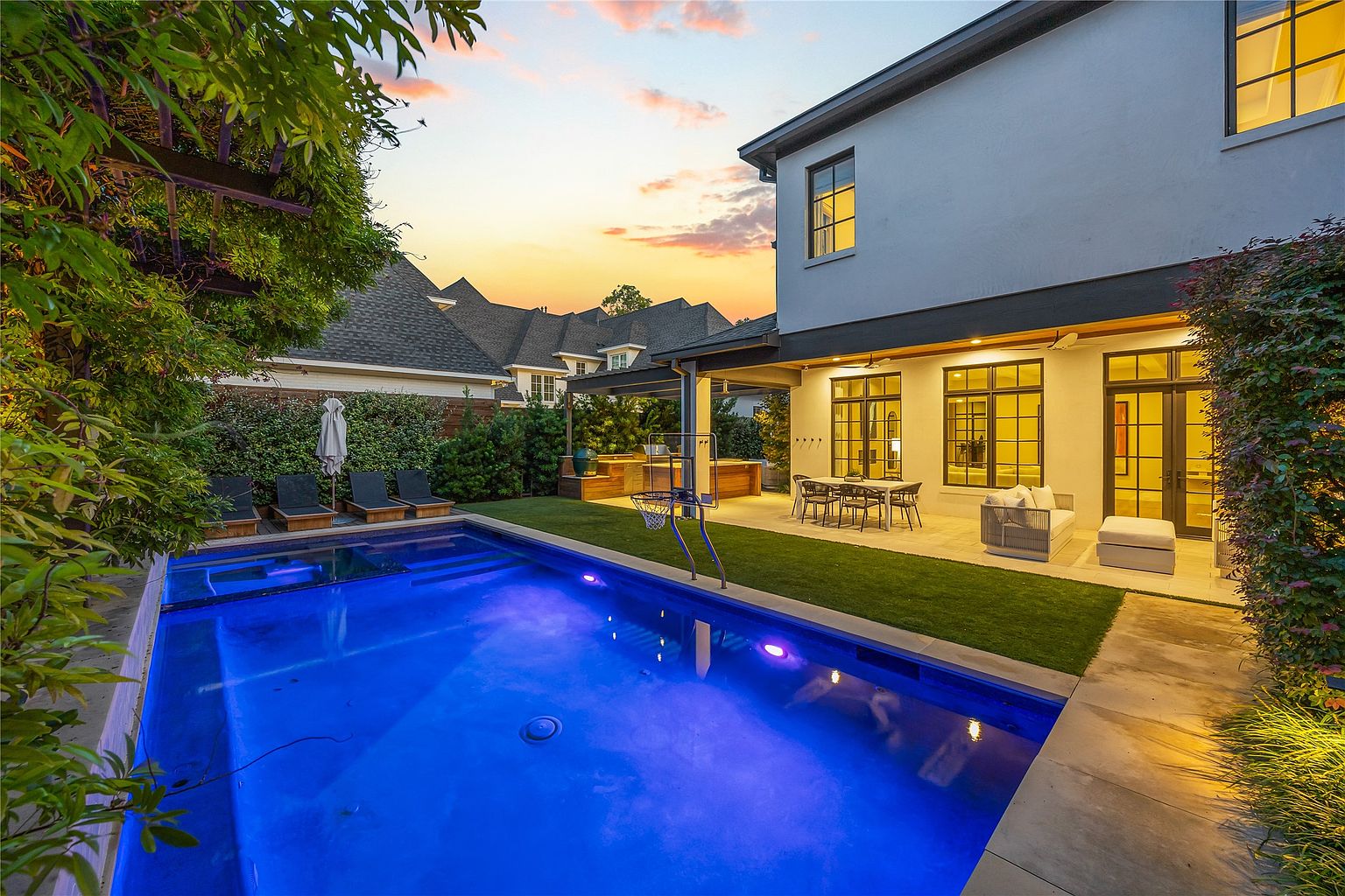 This image showcases a luxurious backyard featuring a modern pool with vibrant blue lighting, surrounded by lush greenery and a well-manicured lawn. The patio area includes comfortable seating and a dining set, perfect for outdoor entertaining. The architecture of the house is contemporary, with large windows and a sleek design, creating an inviting and upscale atmosphere.