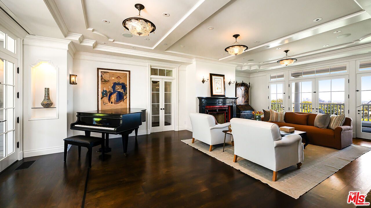 This elegant living room features dark hardwood flooring, a grand piano, and a sophisticated seating area centered around a fireplace with a dark mantel. The space is illuminated by multiple decorative chandeliers and large windows that provide ample natural light and views of the outdoors. The room is decorated with classic artwork and neutral-toned furniture, creating a refined and welcoming atmosphere.