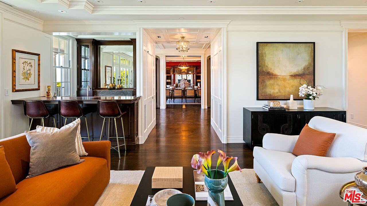 This elegant living area features a warm, inviting color palette with a vibrant orange sofa and a crisp white armchair. The space flows seamlessly into a bar area and a formal dining room visible through a central hallway, showcasing a sophisticated open-concept layout with dark wood flooring. A large abstract painting hangs above a dark console table, adding a touch of modern artistry to the traditional architectural details.
