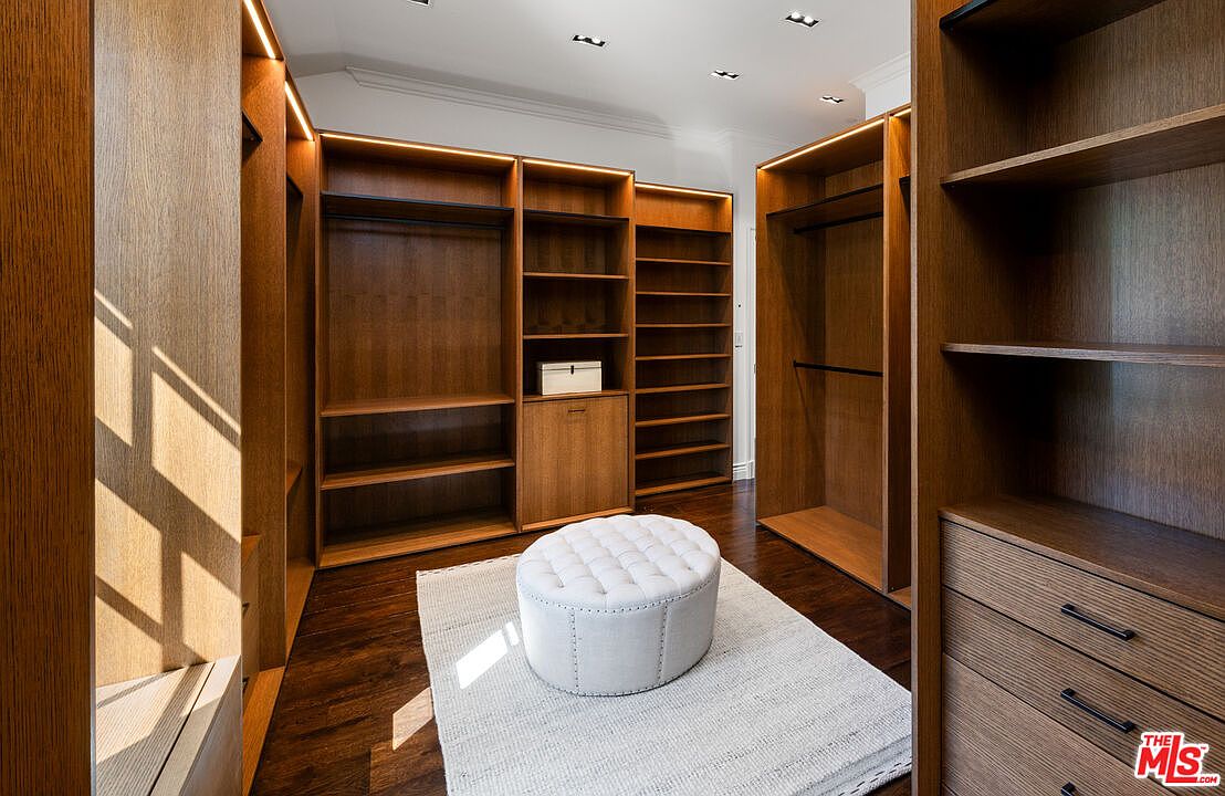 This spacious walk-in closet features custom dark wood cabinetry with integrated LED lighting, providing ample storage and a sophisticated aesthetic. A central tufted ottoman sits atop a light-colored area rug, creating a refined focal point within the room. The dark wood flooring and clean lines contribute to a luxurious and organized atmosphere.