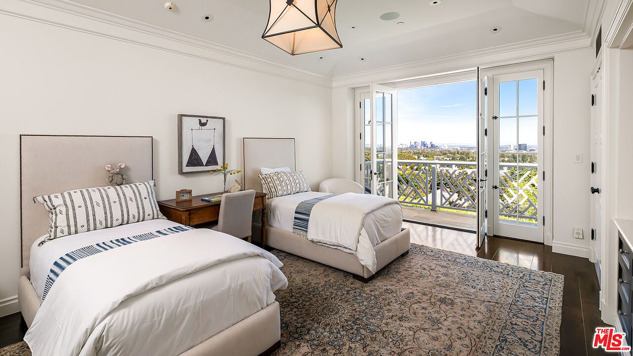 This bright and airy bedroom features two twin beds with neutral upholstered headboards, positioned on either side of a small wooden desk. A large patterned area rug anchors the space, while a geometric pendant light hangs from the ceiling. The room opens through French doors onto a balcony that provides a clear, elevated view of the city skyline.