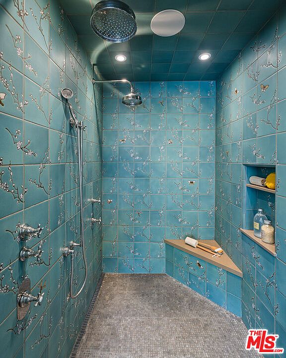 This luxurious walk-in shower features striking teal tiles adorned with a delicate floral and butterfly pattern that extends across the walls and ceiling. The space includes a built-in corner bench, a rainfall showerhead, a handheld sprayer, and a recessed wall niche for storage. A textured stone floor provides a grounding contrast to the vibrant, patterned wall tiles, creating a sophisticated and immersive spa-like atmosphere.
