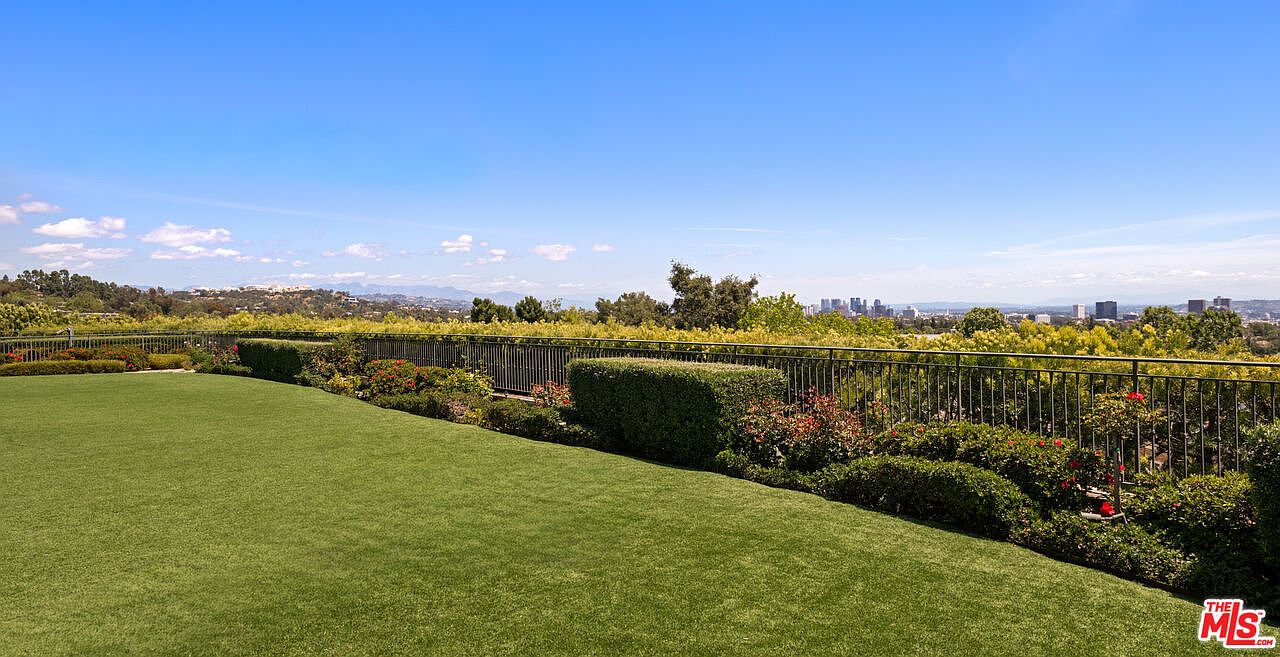 This expansive, manicured lawn offers a serene outdoor space bordered by a metal fence and lush, sculpted hedges. The elevated perspective provides a sweeping, panoramic view of the distant city skyline under a clear blue sky. It is an ideal setting for outdoor entertaining, combining well-maintained landscaping with a dramatic urban backdrop.