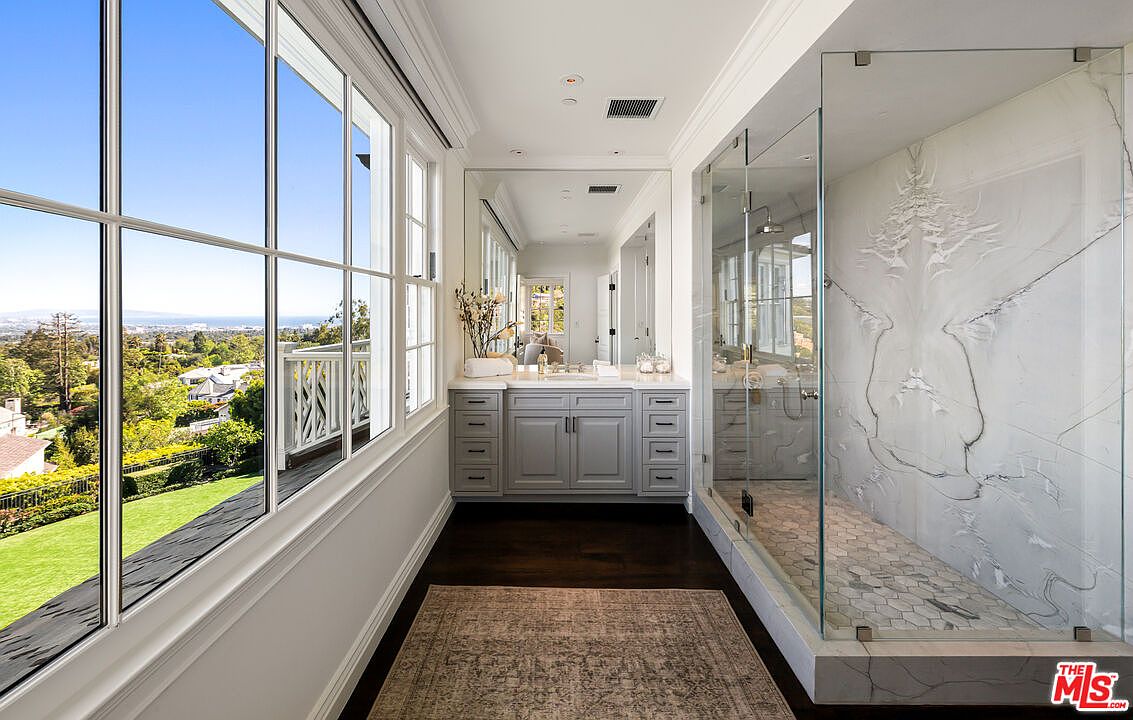 This bright and airy primary bathroom features a large glass-enclosed shower with patterned stone wall tiles and a hexagonal tile floor. A grey vanity with a light-colored countertop sits beneath a large mirror, reflecting the space and the natural light streaming in from the expansive windows. The room is finished with dark wood flooring and a patterned area rug, offering a sophisticated and serene atmosphere with a scenic outdoor view.
