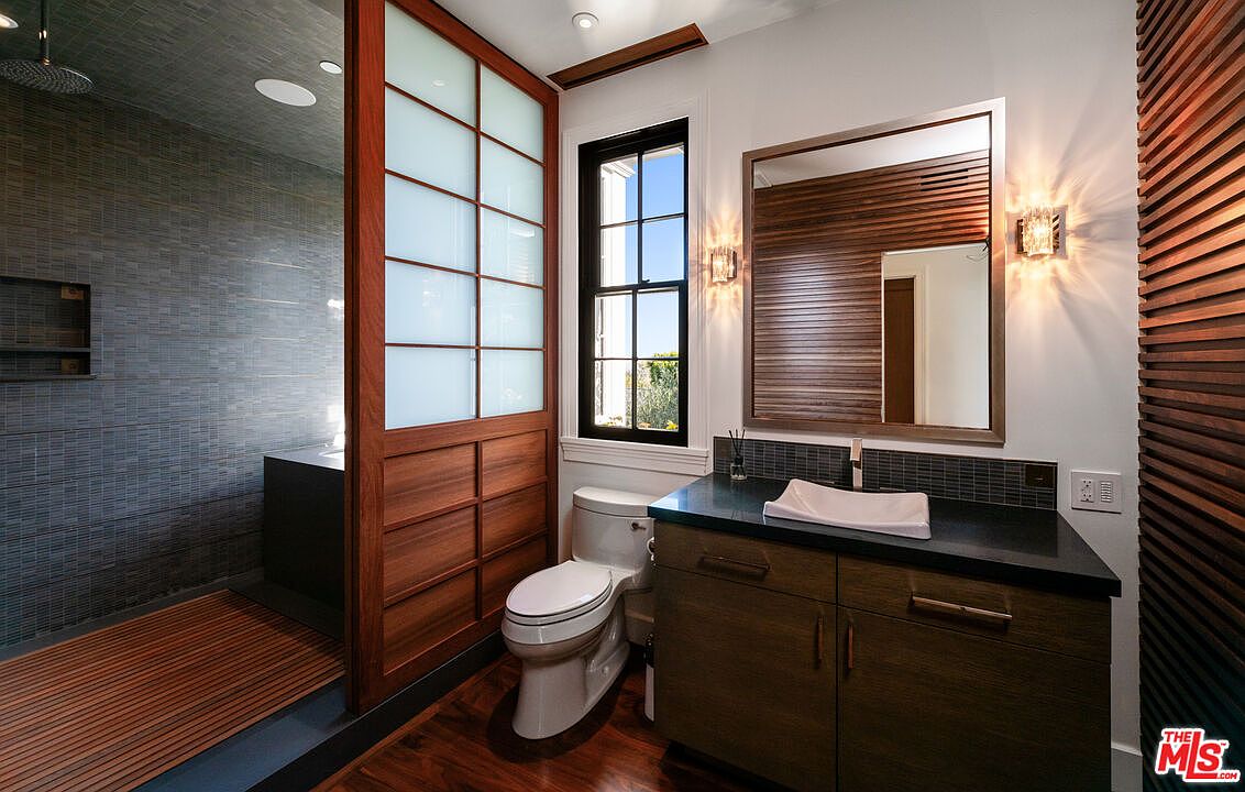 This modern bathroom features a unique Japanese-inspired aesthetic with a shoji-style screen separating the shower area from the vanity. The space includes a dark stone-topped vanity with a vessel sink, a wall-mounted mirror, and a toilet positioned near a window. The combination of warm wood tones and dark, textured wall surfaces creates a sophisticated and tranquil atmosphere.