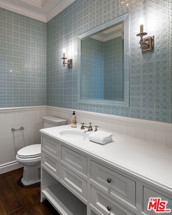 This elegant powder room features a white vanity with a stone countertop and a classic undermount sink. The walls are adorned with light blue patterned tile above a white wainscoting, complemented by two wall-mounted sconces flanking a framed mirror. The space is finished with dark wood flooring and a standard toilet, creating a refined and cohesive aesthetic.