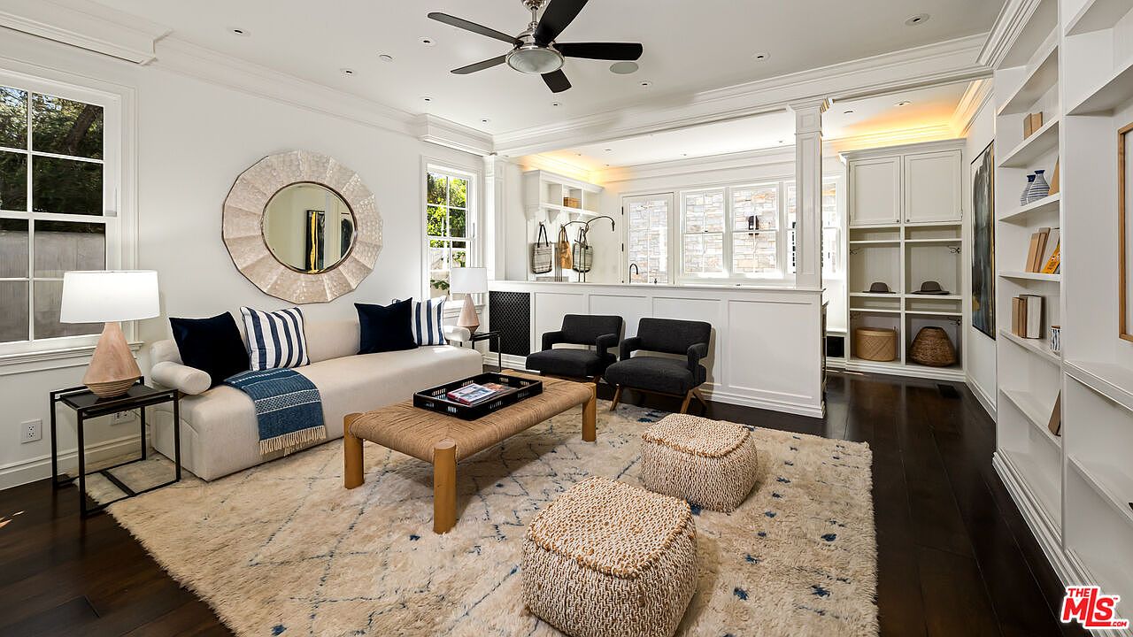 This bright and inviting living room features a neutral color palette with dark wood flooring and a large, textured area rug. The space is furnished with a cream-colored sofa accented by navy pillows, two dark armchairs, and a low-profile coffee table, all centered under a ceiling fan. A large, circular mirror serves as a focal point above the sofa, while built-in shelving units provide ample storage and display space along the side wall.