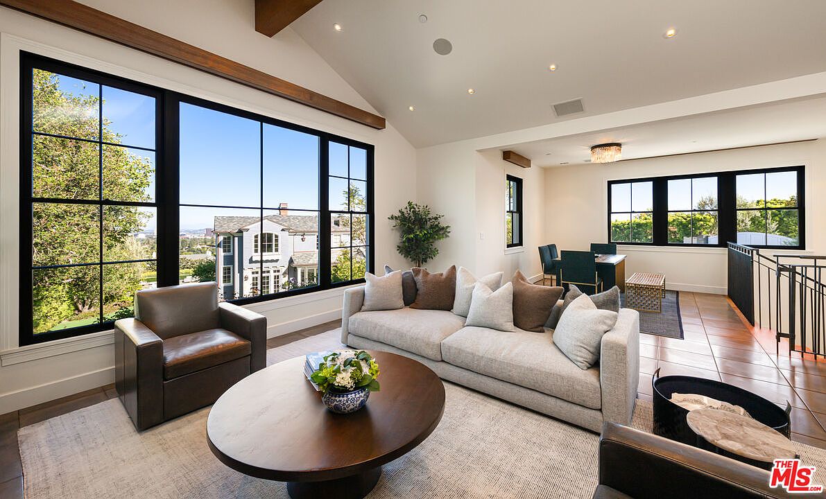 This bright and airy living room features a vaulted ceiling with exposed wood beams and large windows that provide abundant natural light and views of the surrounding neighborhood. The space is furnished with a neutral-toned sofa, a dark leather armchair, and a prominent round wooden coffee table, all set upon a textured area rug. An open-concept layout transitions into a dining area in the background, creating a cohesive and spacious atmosphere suitable for entertaining.