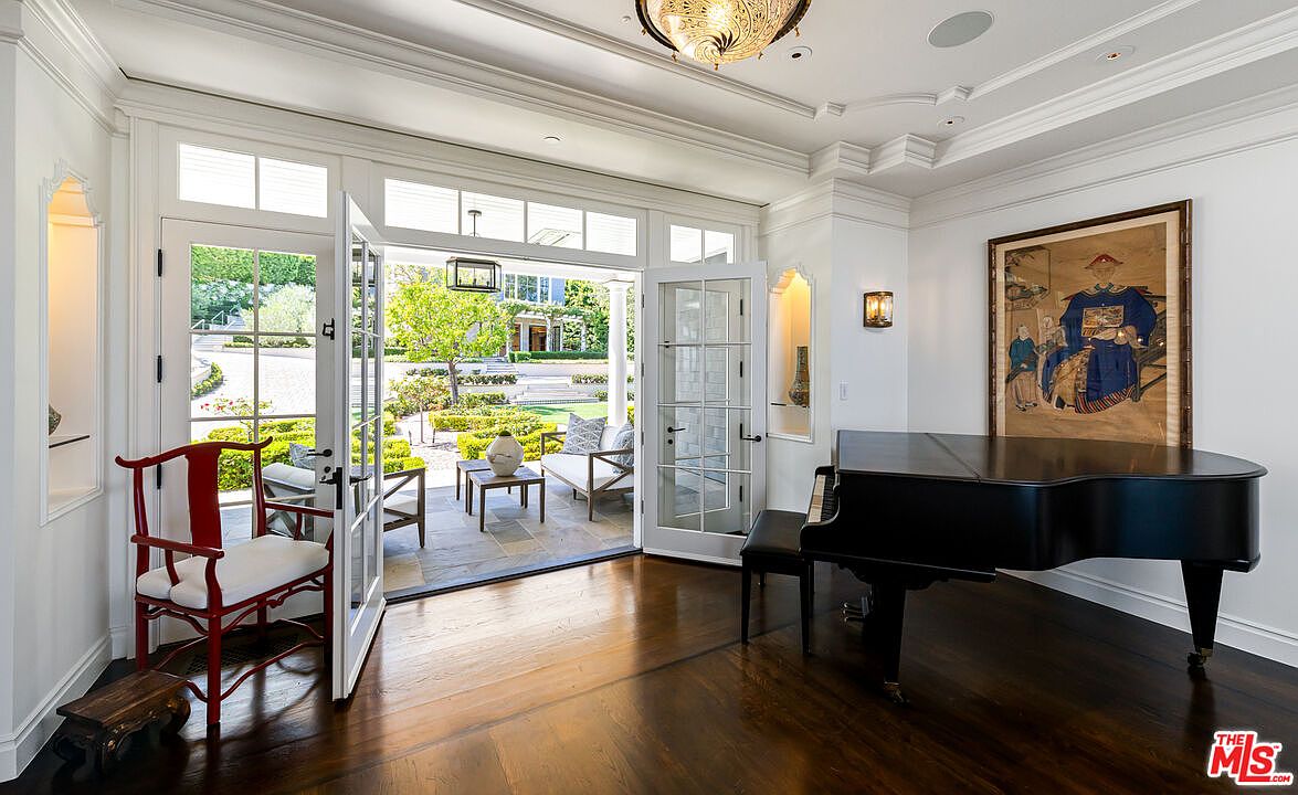 This elegant interior space features dark wood flooring and a grand piano positioned near a large framed portrait. Double glass doors open wide to reveal a sunlit patio and a manicured garden, creating a seamless transition between indoor and outdoor living. The room is accented with a red wooden chair and a decorative ceiling light fixture, conveying a sophisticated and airy atmosphere.