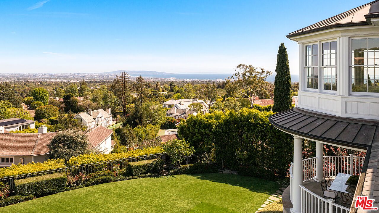 This elevated rear view captures a sprawling, manicured lawn bordered by lush greenery and a metal fence, overlooking a vast suburban landscape that extends toward the distant ocean. To the right, a multi-story home features a classic architectural style with a wrap-around porch, white siding, and large windows that offer expansive views. The perspective is cinematic, emphasizing the property's prime location and the seamless transition between the private outdoor space and the scenic horizon.