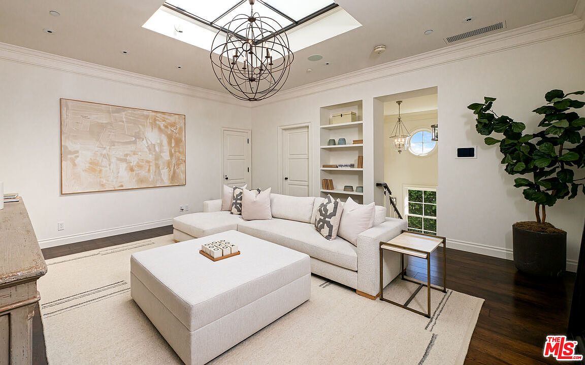 This bright and airy living room features a large white sectional sofa paired with a matching oversized ottoman, creating a comfortable seating area. A striking spherical chandelier hangs from the center of the room beneath a large skylight, while built-in shelving and a large abstract art piece add character to the space. The room is finished with dark wood flooring and a neutral area rug, providing a sophisticated and inviting atmosphere.