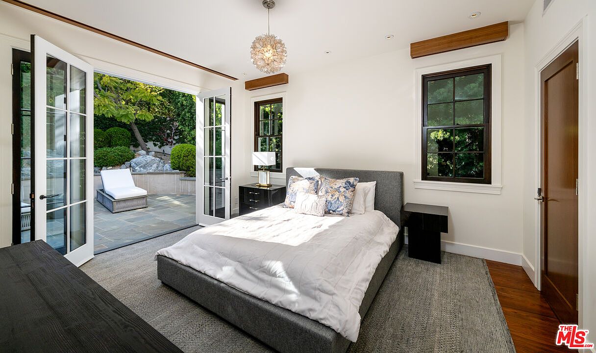 This bright and airy bedroom features a neutral color palette and direct access to a private stone-paved patio through large glass-paned double doors. The room is anchored by a gray upholstered bed with patterned accent pillows, flanked by dark bedside tables and a modern chandelier. The space blends indoor comfort with outdoor living, creating a serene and inviting atmosphere.