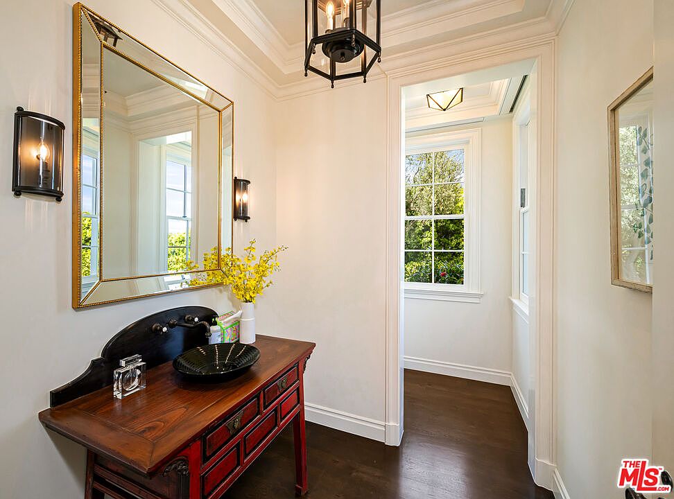 This elegant powder room features a dark wood vanity with a unique black vessel sink and wall-mounted faucet. A large, geometric-shaped mirror with a gold frame hangs above the vanity, flanked by two black wall sconces, while a bright yellow floral arrangement adds a pop of color. The space leads into a sunlit hallway with dark wood flooring and a window looking out onto greenery.