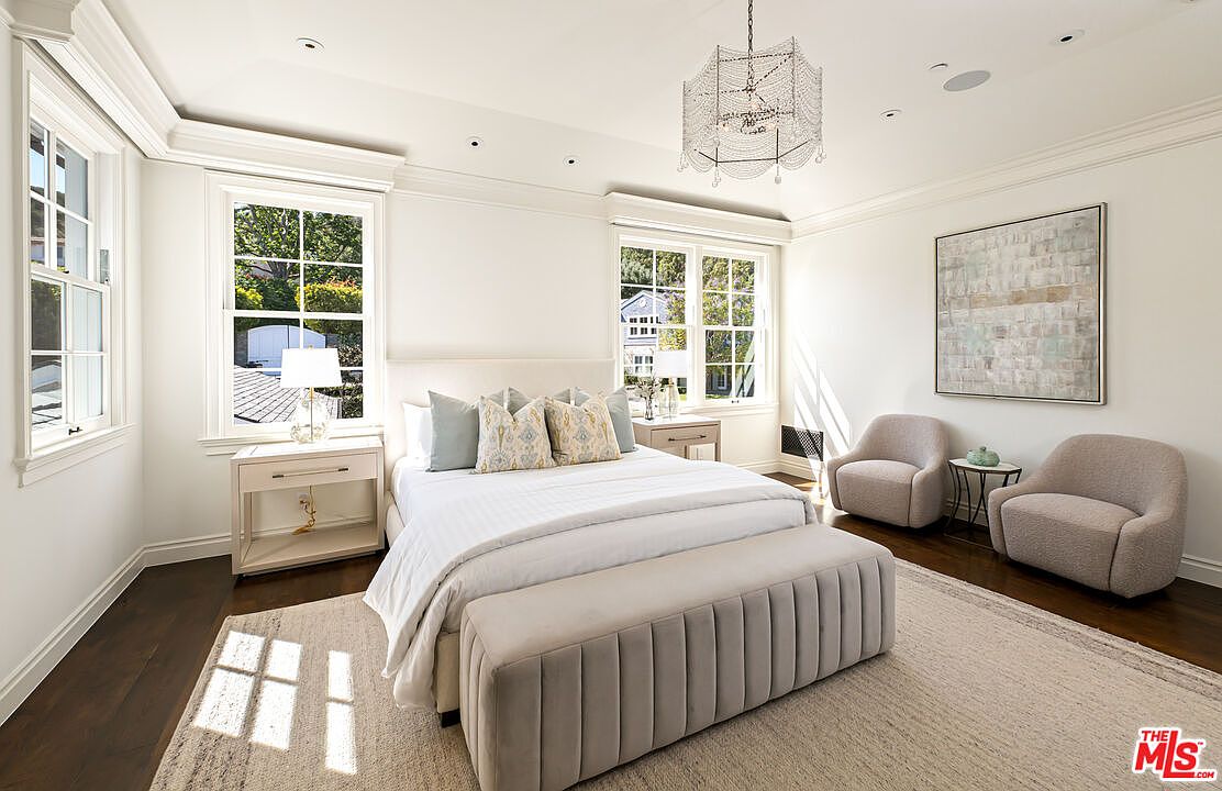 This bright and airy primary bedroom features a neutral color palette with soft textures and elegant crown molding. A plush, tufted bench sits at the foot of the bed, while two comfortable armchairs create a cozy seating area near a large piece of abstract wall art. The room is illuminated by a decorative chandelier and natural light streaming through multiple windows, creating a serene and sophisticated atmosphere.