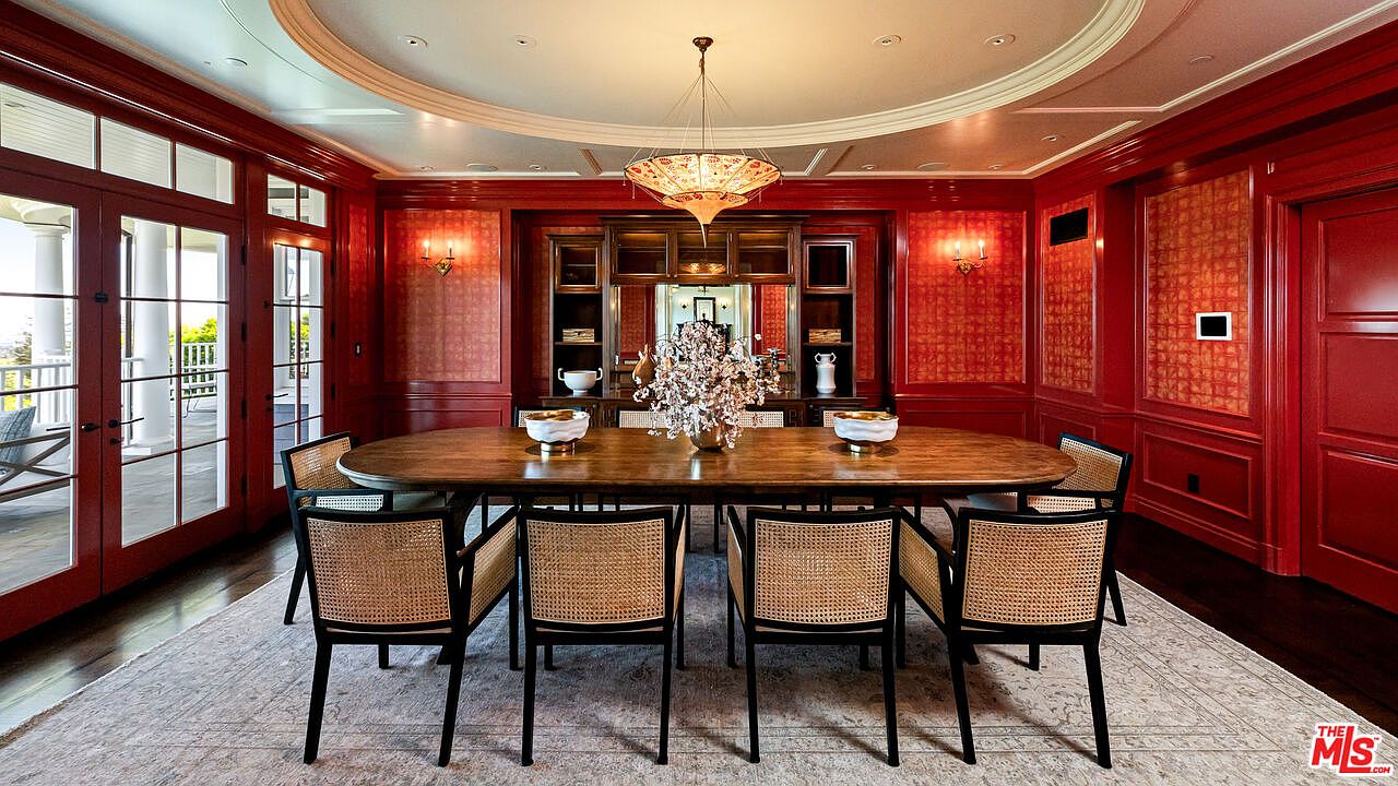 This formal dining room features bold red paneled walls and a large, oval wooden table surrounded by six chairs with woven cane backs. A decorative chandelier hangs from the center of a recessed ceiling, while a built-in hutch with glass-front cabinets provides an elegant display area. The room is anchored by a large, neutral-toned area rug and offers direct access to an outdoor space through tall glass doors.