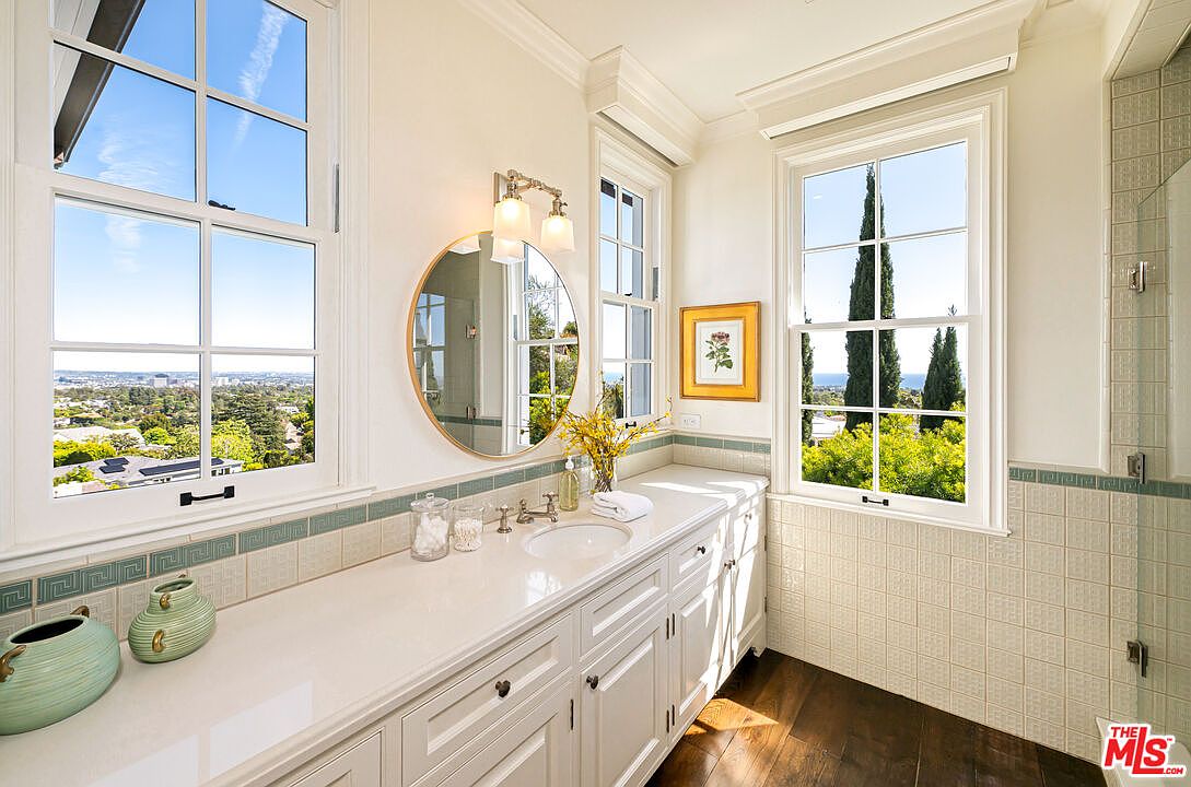 This bright and airy bathroom features a long white vanity with a light-colored countertop, a circular mirror, and dual wall-mounted light fixtures. Large windows on either side of the vanity provide ample natural light and expansive views of the surrounding landscape. The space is finished with light-toned wall tiles and dark wood flooring, creating a clean and elegant aesthetic.