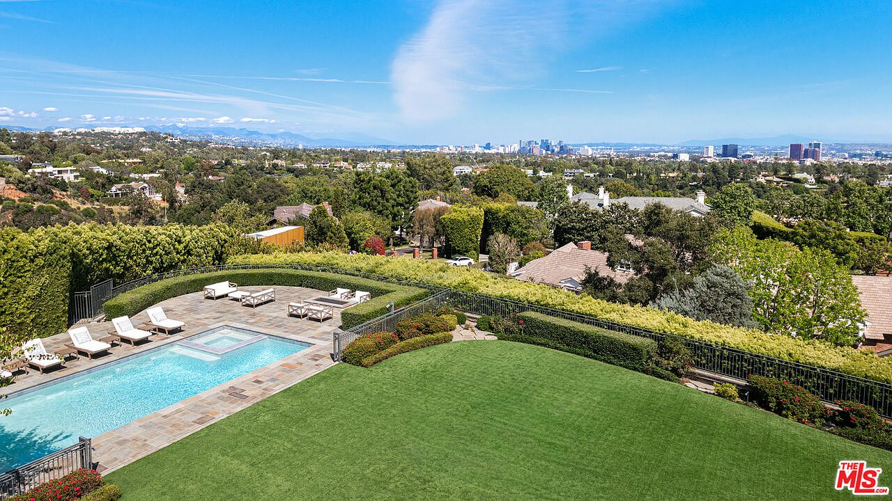 This elevated perspective captures a sprawling backyard featuring a rectangular swimming pool with an integrated spa and a stone-paved patio area furnished with lounge chairs. A lush, manicured lawn stretches out toward a black metal fence, beyond which lies a dense canopy of trees and a distant cityscape under a clear blue sky. The scene conveys a sense of luxury and privacy, highlighting the property's expansive outdoor living space and scenic views.