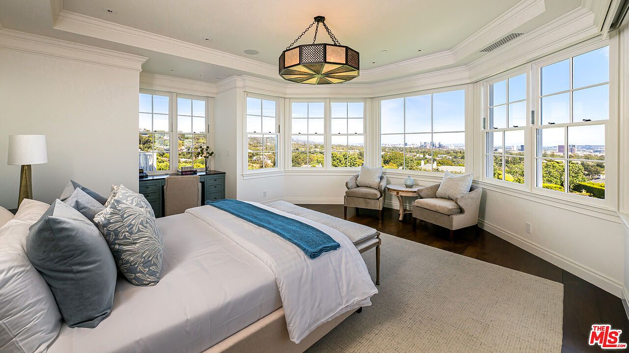 This spacious primary bedroom features a neutral color palette and a large bay window area that offers expansive views of the city skyline. The room is furnished with a plush bed, two comfortable armchairs, and a dedicated desk space, all set against dark wood flooring and a light-colored area rug. A unique, multi-faceted pendant light fixture serves as a focal point, enhancing the sophisticated and airy atmosphere of the space.