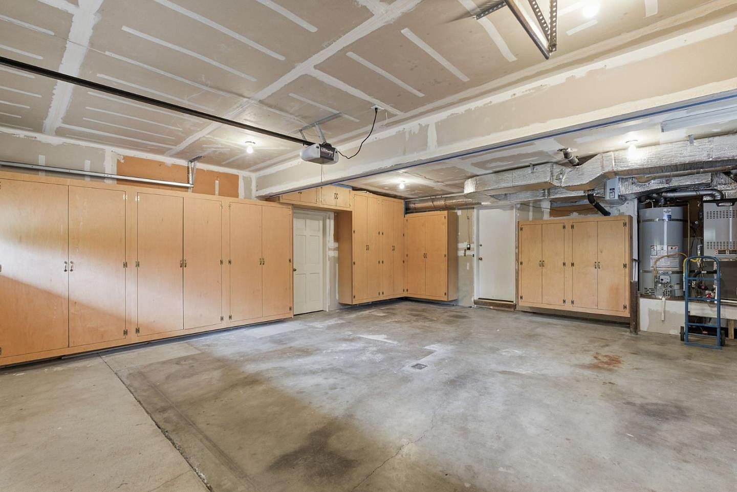 This is an interior shot of a spacious garage. The garage features built-in wooden cabinets along the walls for ample storage. The concrete floor is expansive, and the ceiling is unfinished, showing the structure and some utilities. A water heater is visible in one corner, and there are two doors, one leading to the interior of the house and the other to the outside.
