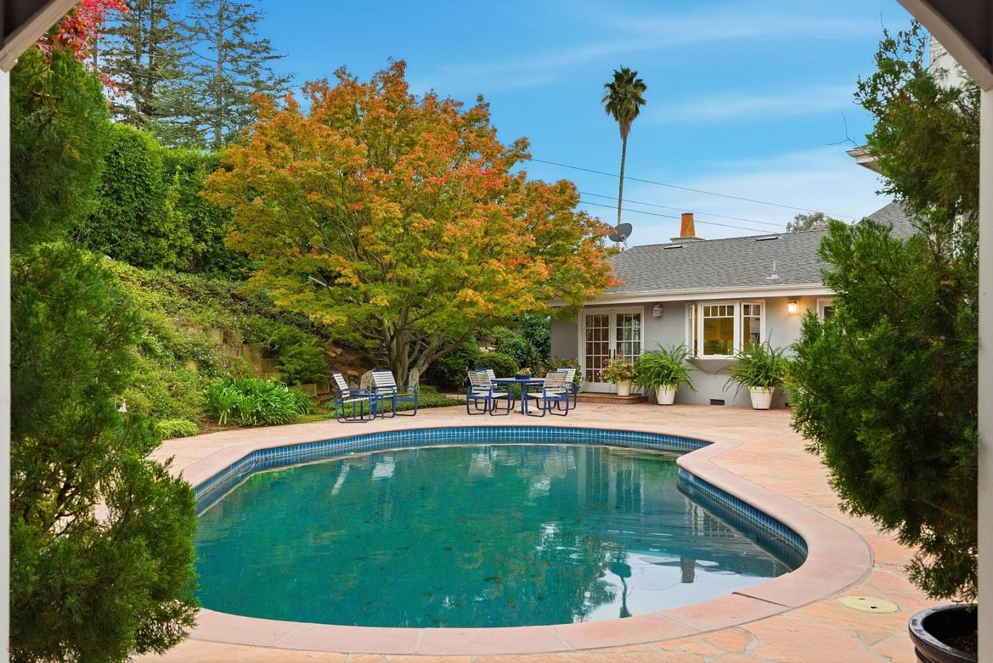 This image showcases a beautiful backyard pool area. The kidney-shaped pool is surrounded by a stone patio and lush landscaping, including mature trees and shrubs. A patio table and chairs are set up near the pool, suggesting a relaxing outdoor living space. The house is visible in the background, with a well-maintained exterior and windows overlooking the pool area.