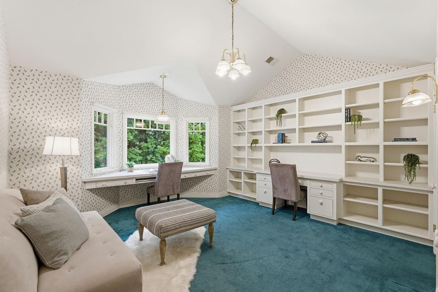 This is an interior shot of a home office or study. The room features built-in bookshelves, a long desk area with two workstations, and a cozy seating area with a sofa and ottoman. The walls are covered in a subtle patterned wallpaper, and the room is lit by a chandelier and a table lamp, creating a warm and inviting atmosphere.