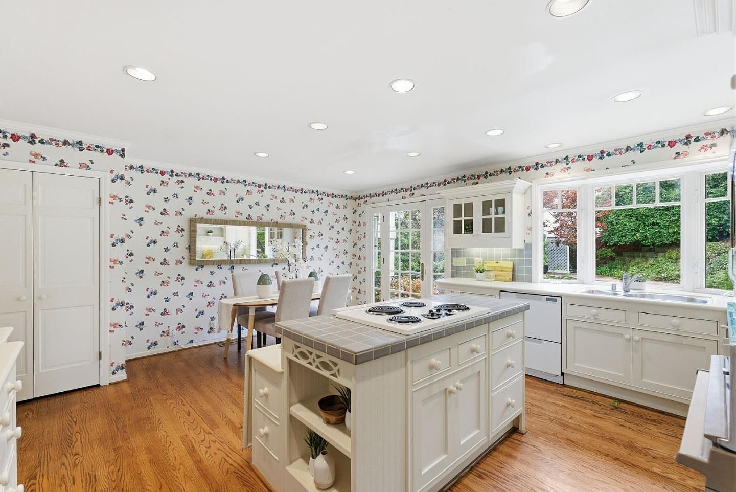 This is a bright and airy kitchen featuring white cabinetry, a central island with a cooktop, and hardwood floors. The walls are adorned with floral wallpaper, adding a touch of vintage charm. A dining area is visible in the background, creating an open and inviting space.