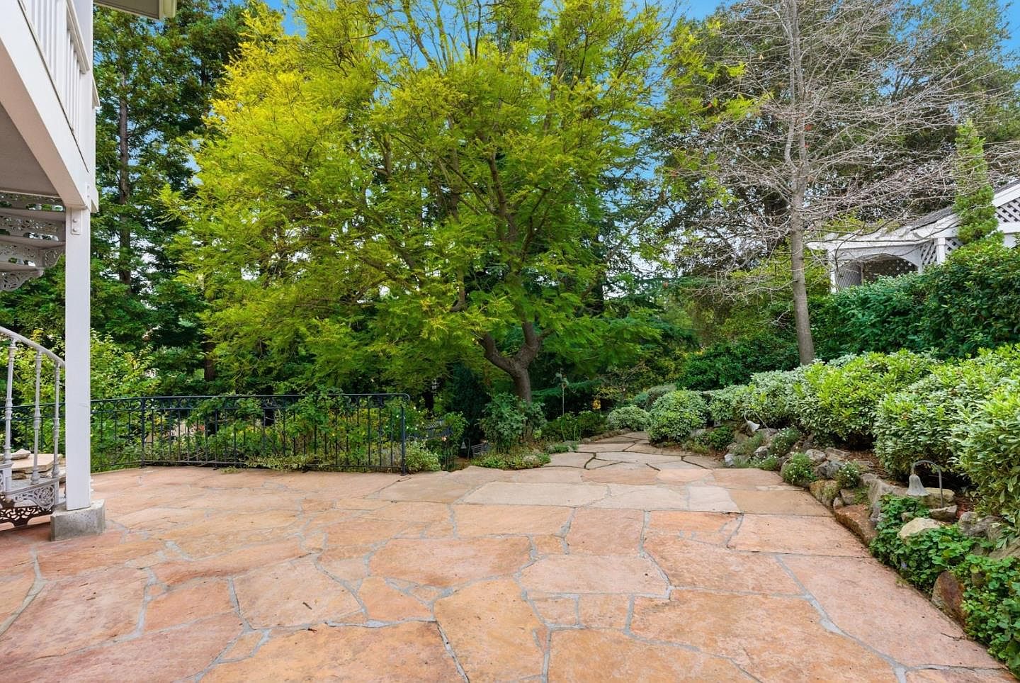 This image showcases a spacious outdoor patio area, featuring flagstone paving and a wrought iron railing. Lush greenery and mature trees surround the patio, creating a private and serene atmosphere. The space appears ideal for outdoor entertaining or relaxation, adding significant value to the property.