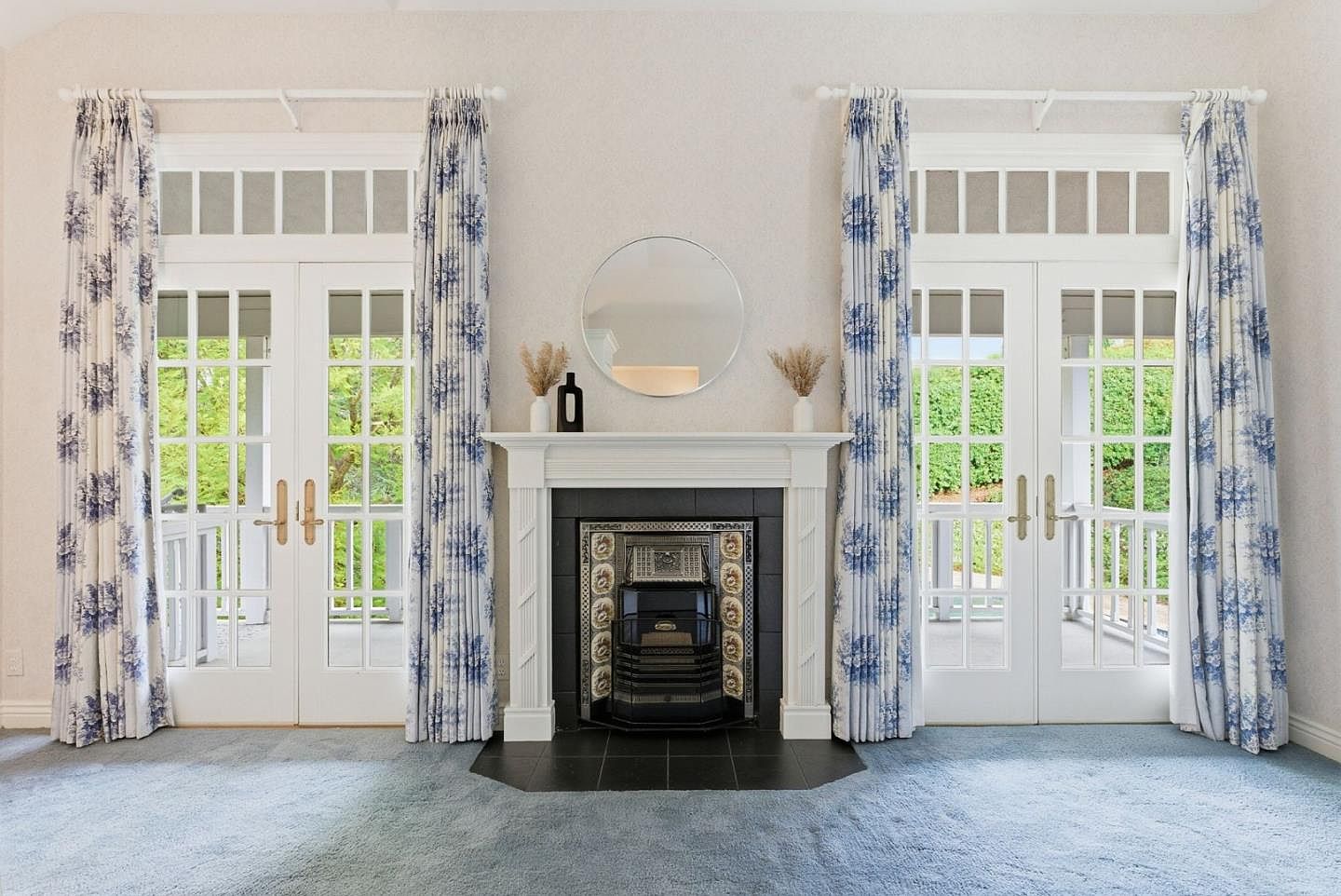This living room features a classic fireplace with a white mantel and a round mirror above. Two sets of French doors with white frames and blue floral curtains flank the fireplace, leading to outdoor spaces. The room is carpeted in a light blue, creating a serene and inviting atmosphere.
