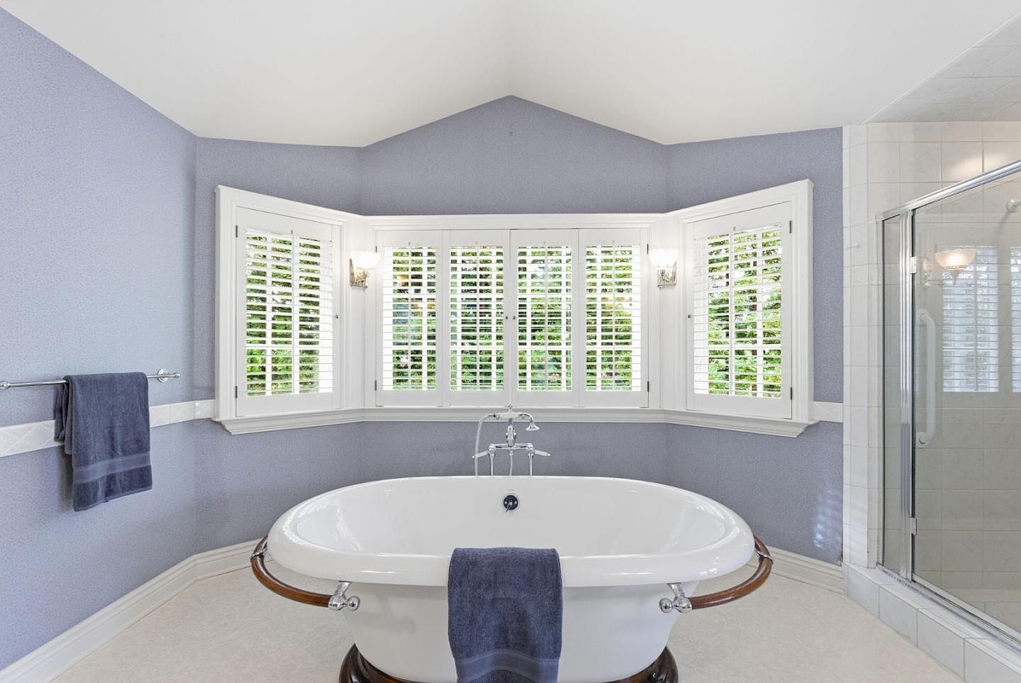 This is a luxurious primary bathroom featuring a freestanding oval bathtub with a wooden accent and chrome fixtures, positioned in front of a bay window with white shutters. The walls are painted in a soft blue hue, and a glass-enclosed shower is visible to the right. The overall impression is one of tranquility and spa-like elegance.