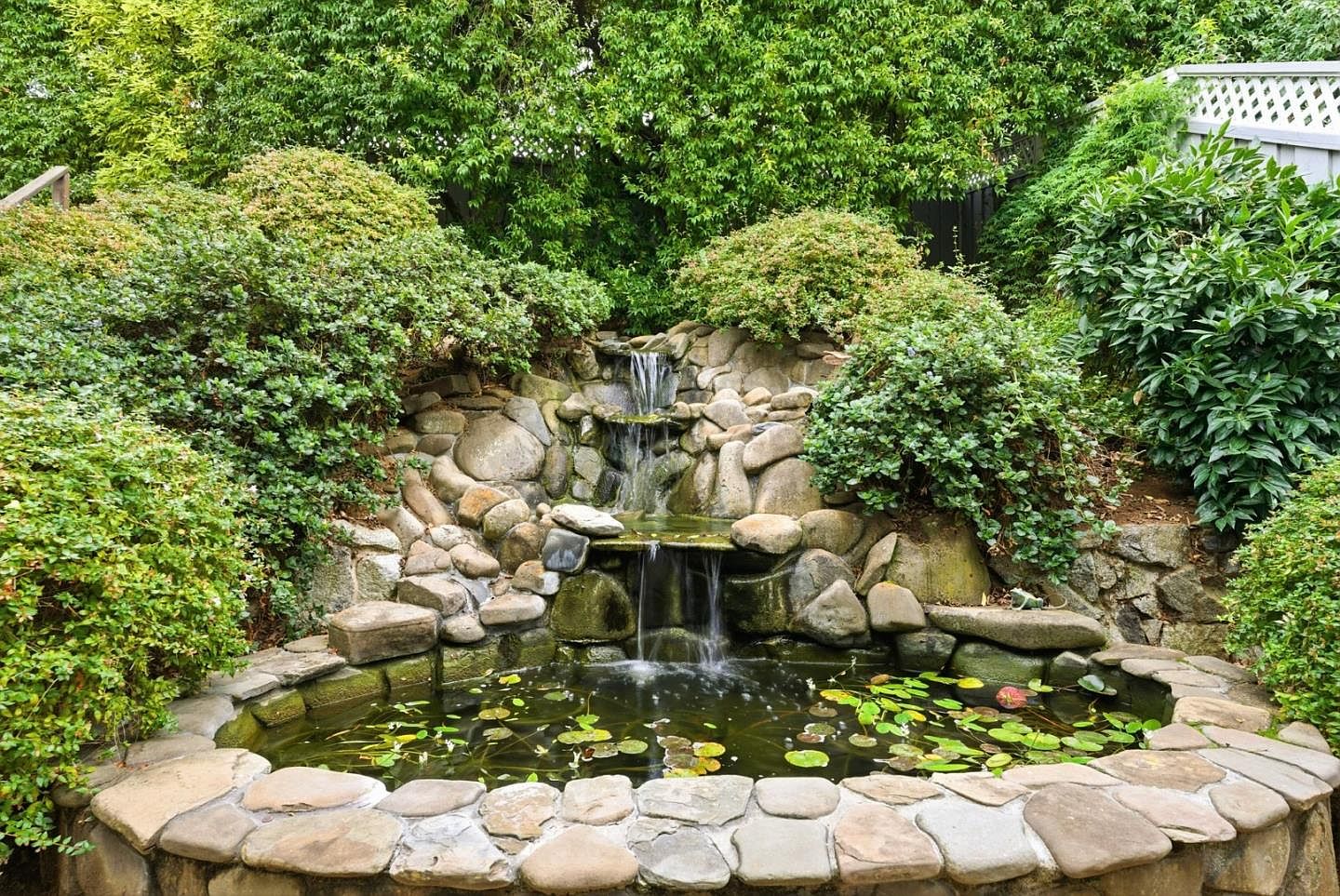 This image showcases a serene backyard garden featuring a multi-tiered rock waterfall that flows into a small pond. The pond is surrounded by a stone border and contains lily pads, creating a tranquil and natural aesthetic. Lush greenery and bushes frame the waterfall, enhancing the garden's peaceful ambiance.