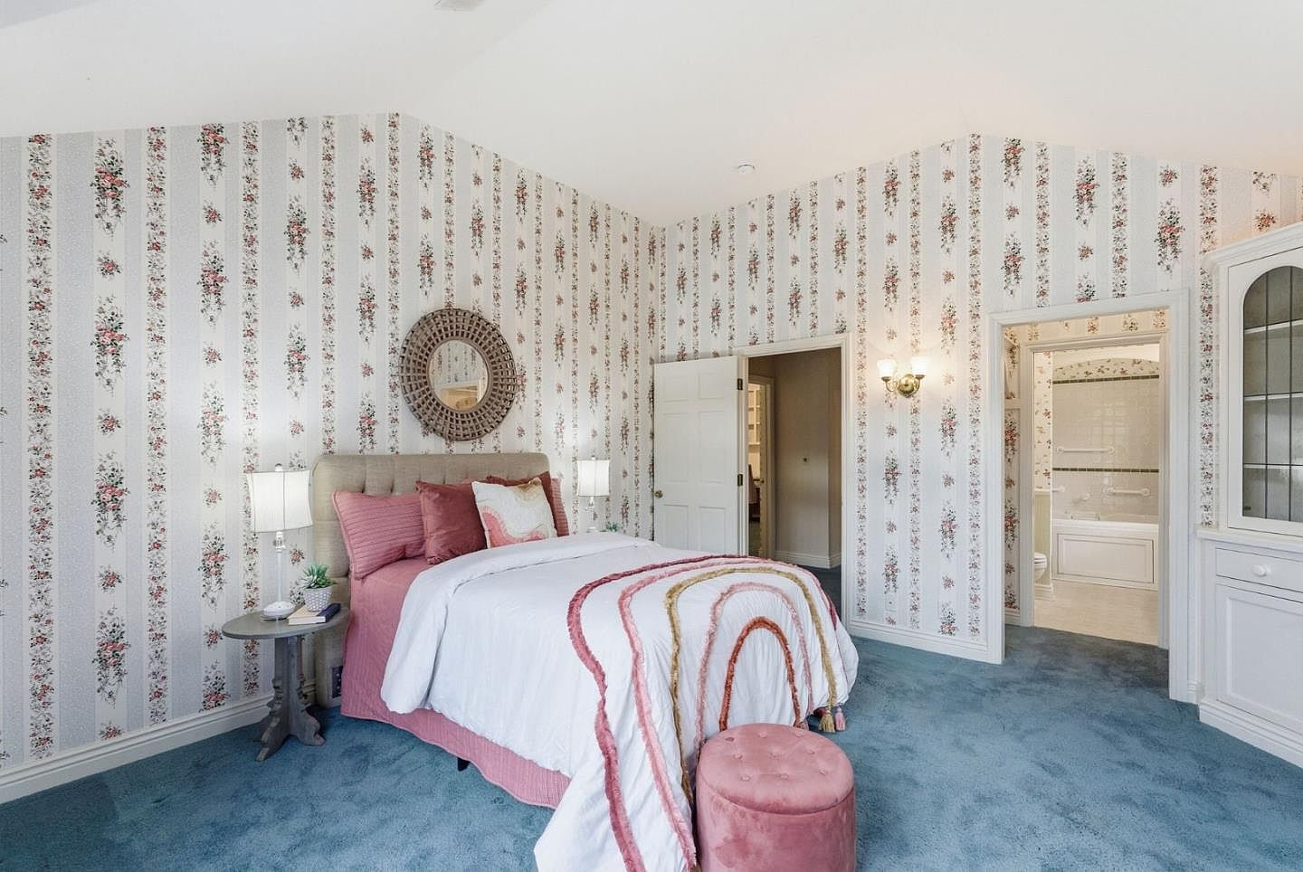 This bedroom features a floral wallpaper, blue carpet, and a bed with pink and white bedding. A round mirror hangs above the bed, and a pink ottoman sits at the foot of the bed. The room has a cozy and vintage aesthetic, with doorways leading to other rooms.