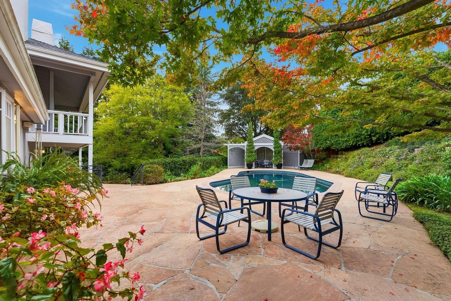 This image showcases a beautifully landscaped backyard featuring a stone patio with outdoor seating, a swimming pool, and a charming gazebo. Mature trees provide shade and privacy, while colorful flowers add a touch of vibrancy. The overall impression is one of tranquility and luxury, perfect for outdoor entertaining and relaxation.
