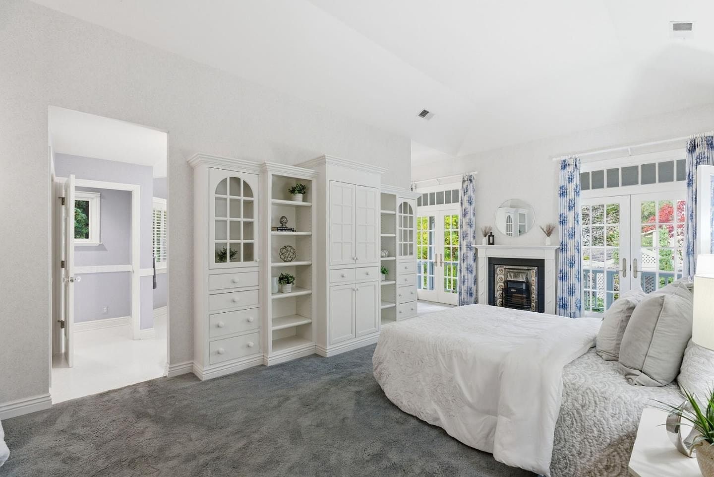 This is a bright and spacious primary bedroom featuring a built-in white shelving unit and a fireplace with French doors leading to the outside. The room is carpeted in a neutral tone, and the bed is neatly made with white linens and decorative pillows. The overall impression is one of comfort and elegance.
